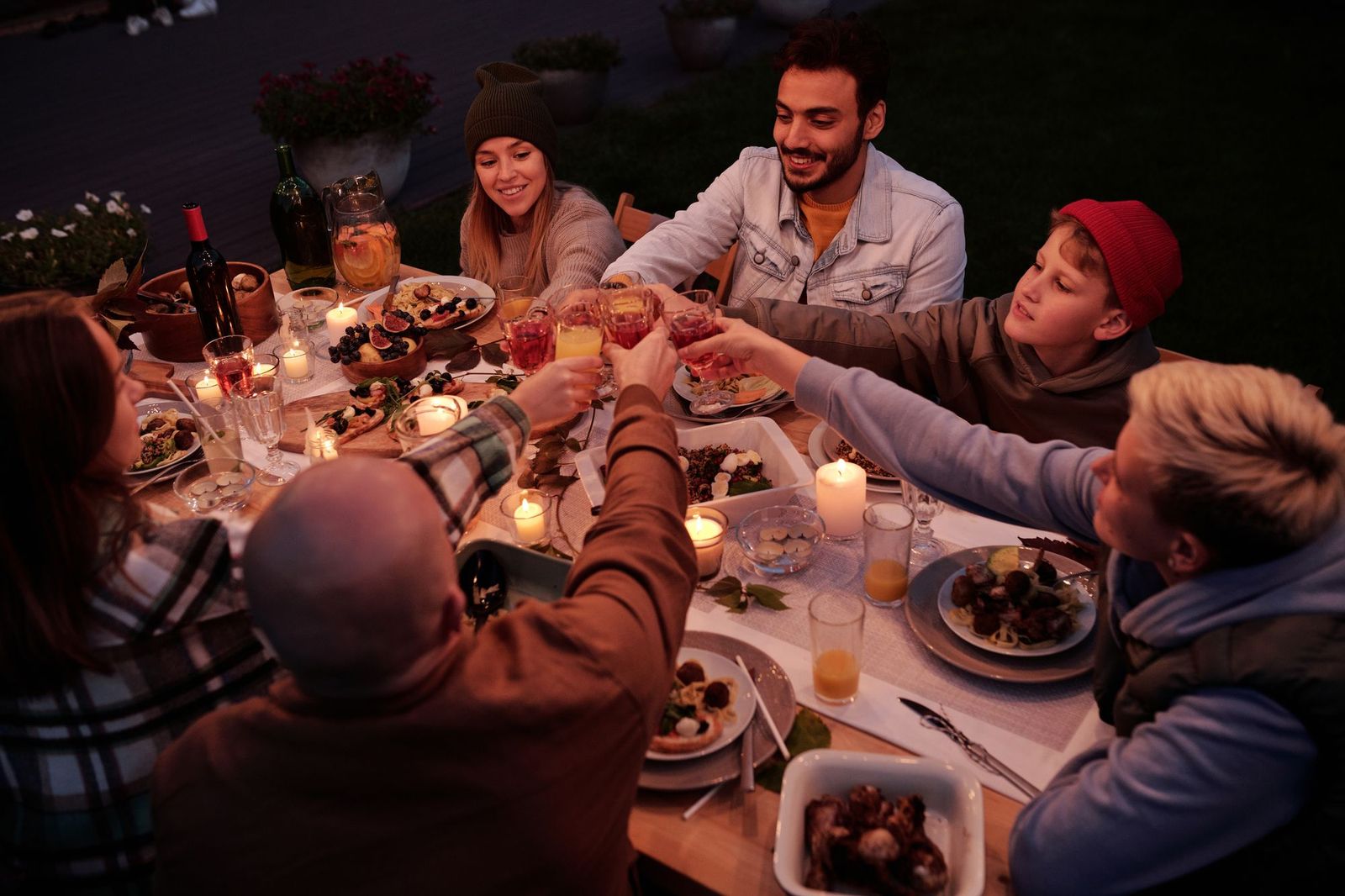 A group of friends sitting around a long candlelit table in a garden at dusk, raising their glasses together